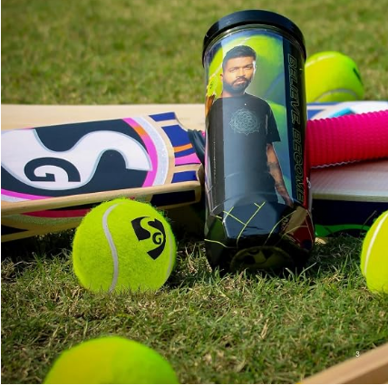 A black cylindrical tennis ball can with a printed portrait of Virat Kohli, standing upright on grass alongside tennis balls and a cricket bat.