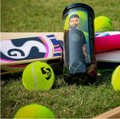 A black cylindrical tennis ball can with a printed portrait of Virat Kohli, standing upright on grass alongside tennis balls and a cricket bat.